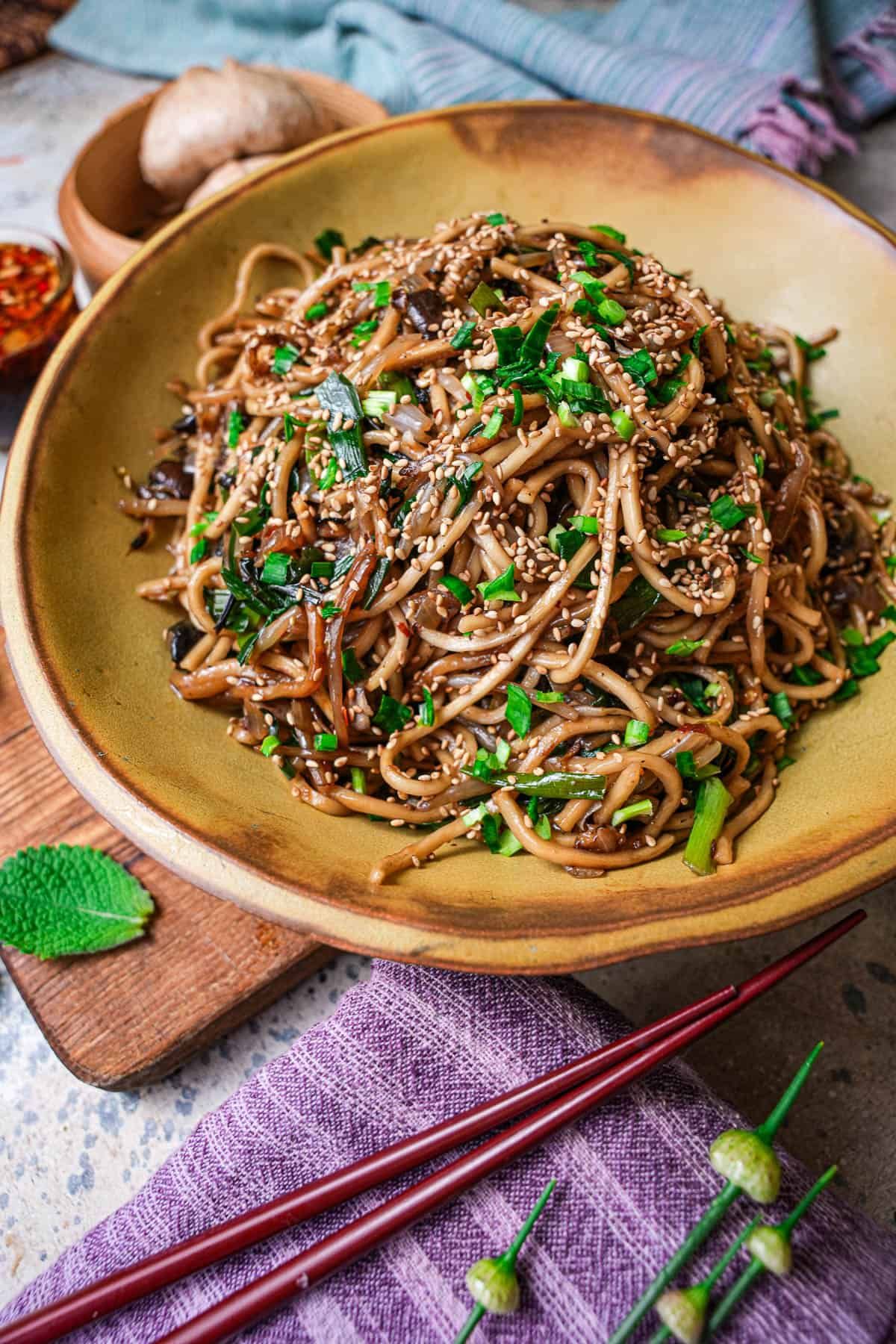 Soba noodles with mushrooms, sesame seeds, and scallions in a brown bowl. Chopsticks and purple cloth.