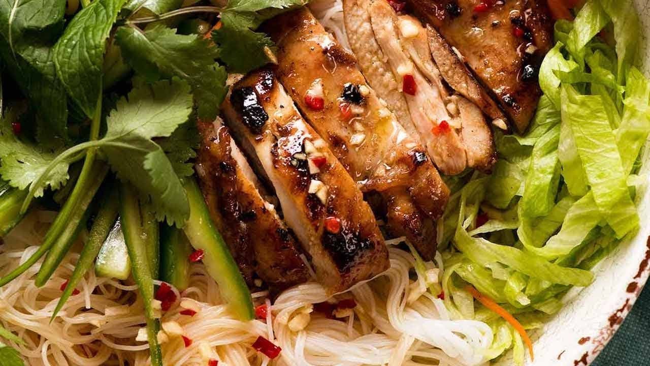 Grilled chicken slices over vermicelli noodles and fresh vegetables in a bowl.