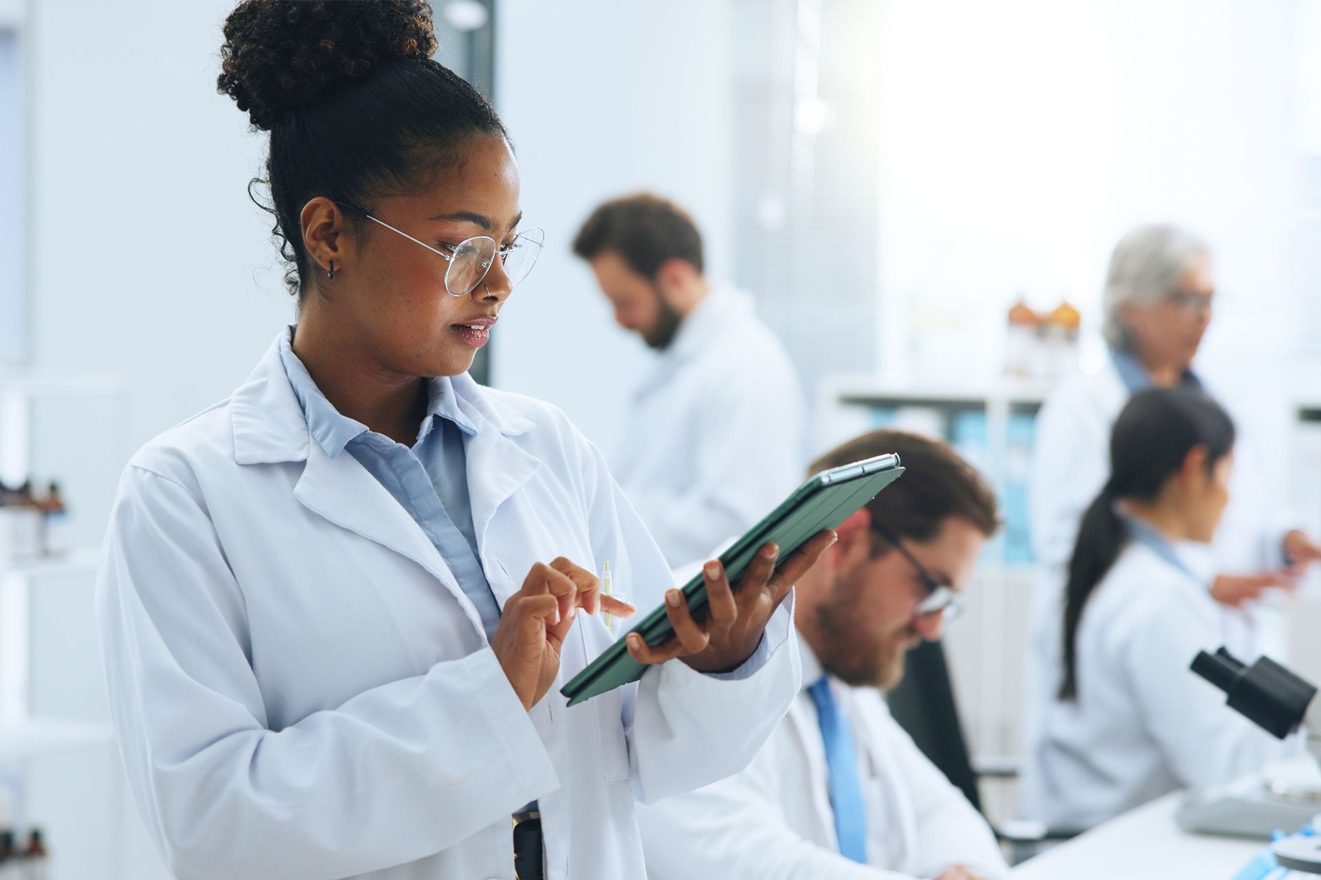 Scientists in white coats work in a bright lab, one using a tablet while others collaborate.