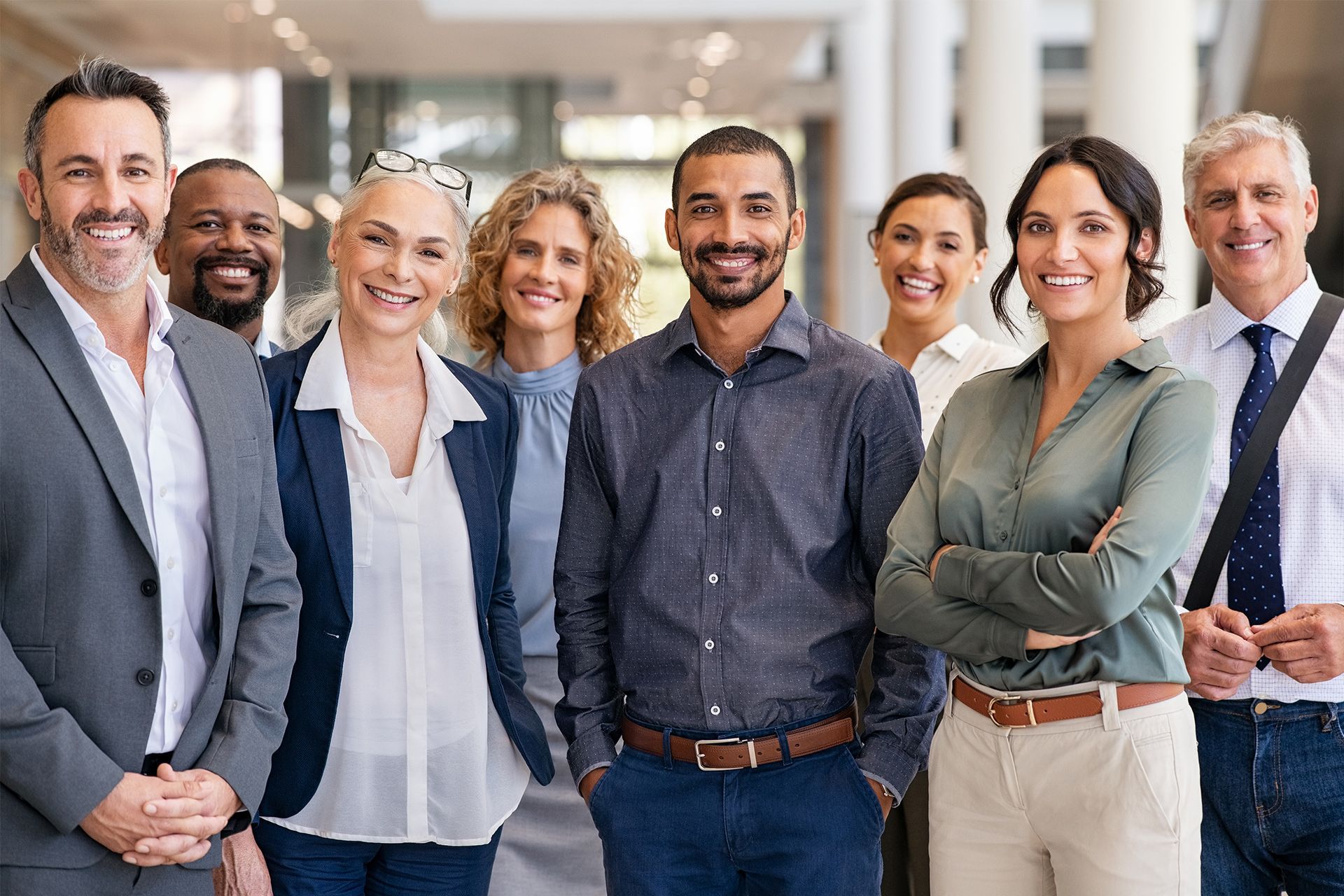Group of eight coworkers smiling in a bright office hallway, standing shoulder to shoulder.