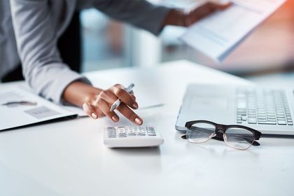 Person counting money with a calculator beside a laptop and eyeglasses on a desk