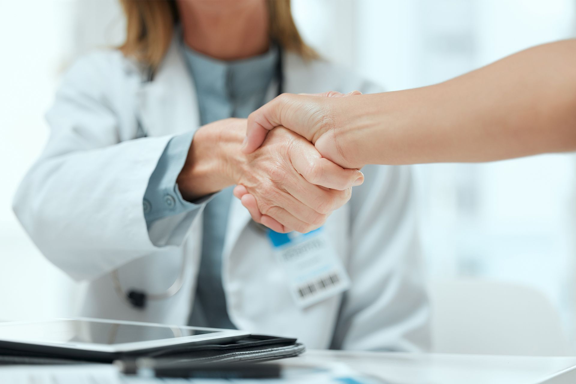 Handshake with a doctor in a white coat across a desk, with a laptop in the foreground.