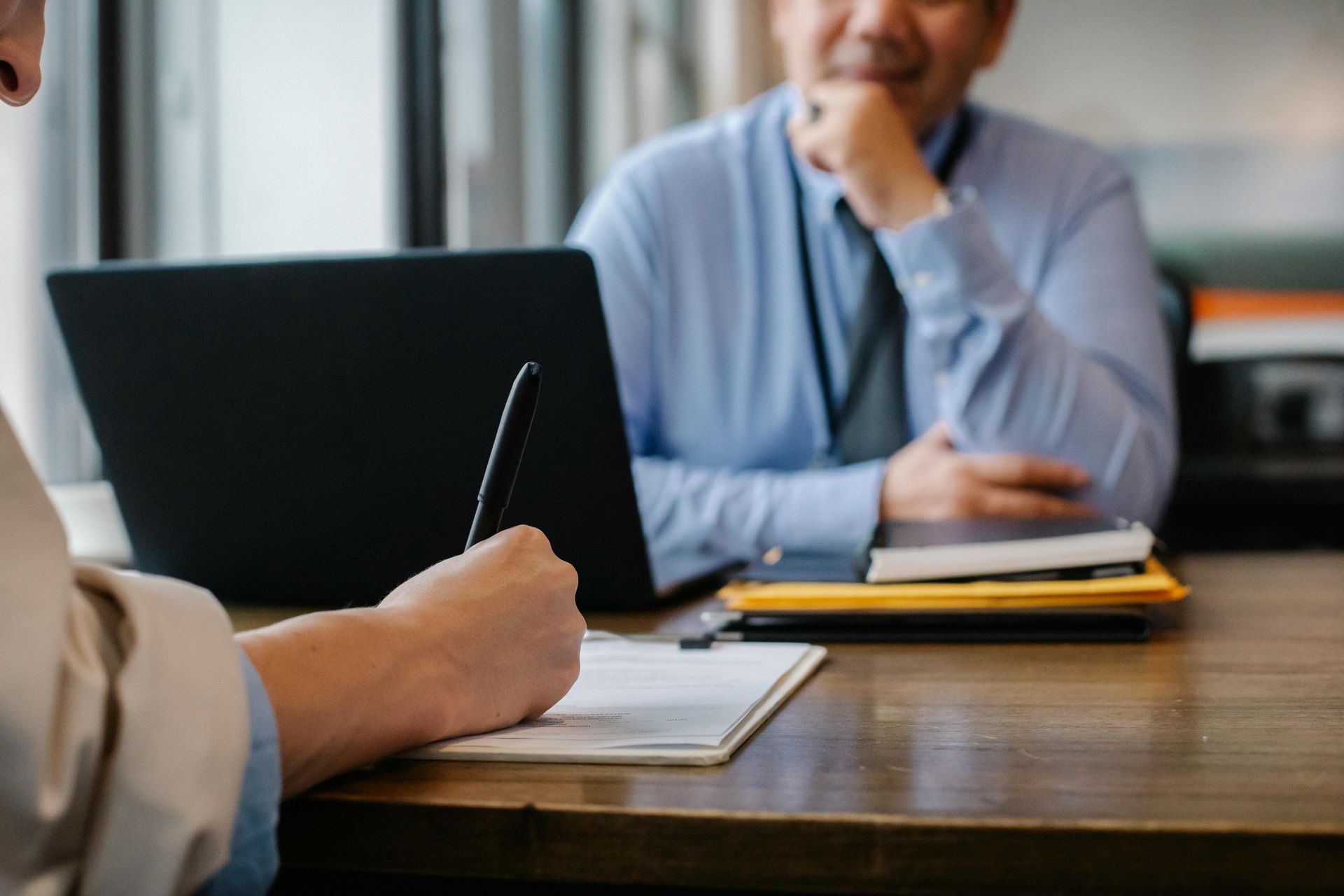 Two people in a meeting taking notes beside a laptop on a wooden table