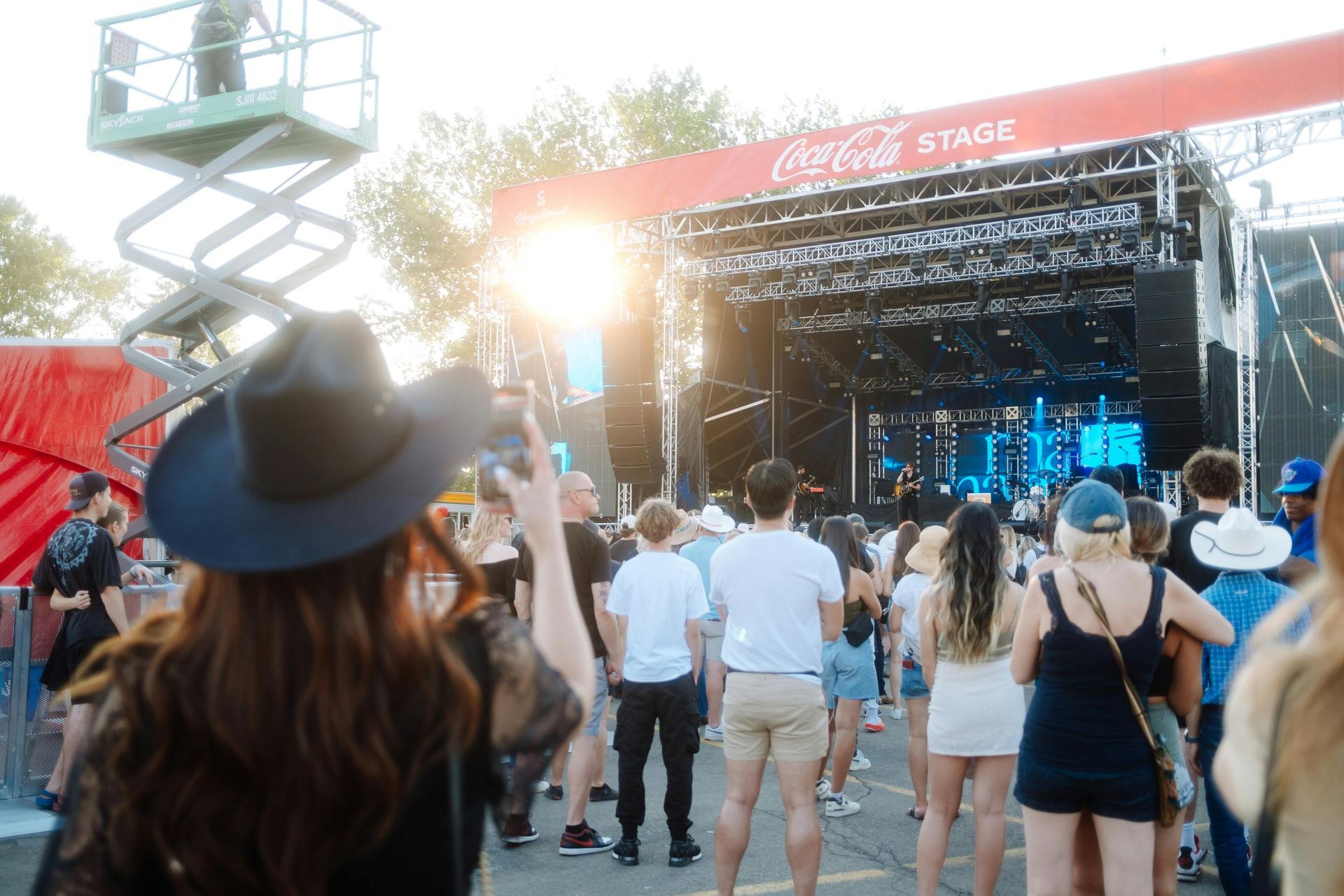 Crowd facing a lit outdoor stage at a music festival, with a person in a black hat in the foreground