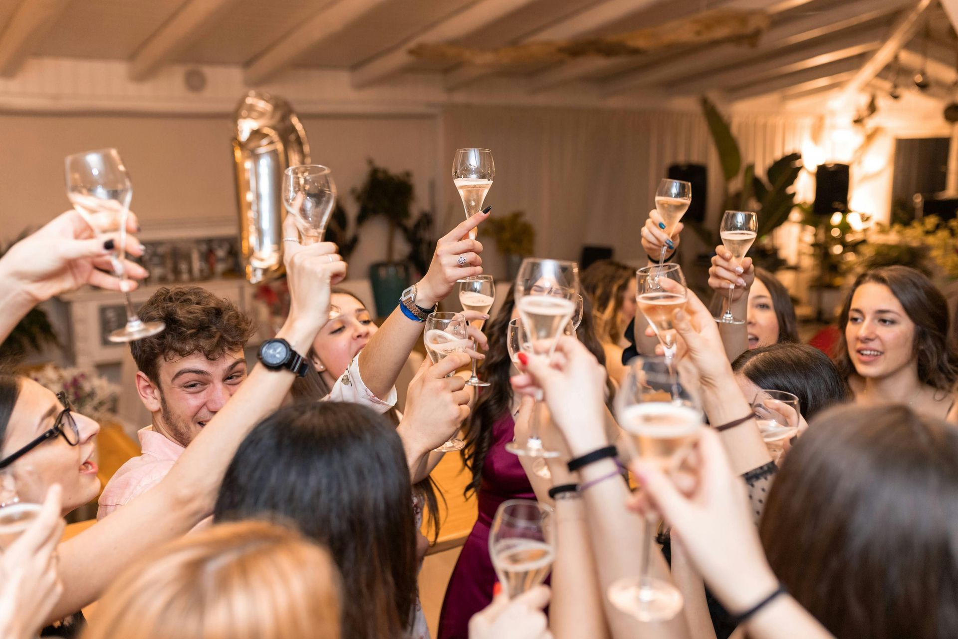 People raising champagne glasses in a lively indoor celebration, smiling and cheering.