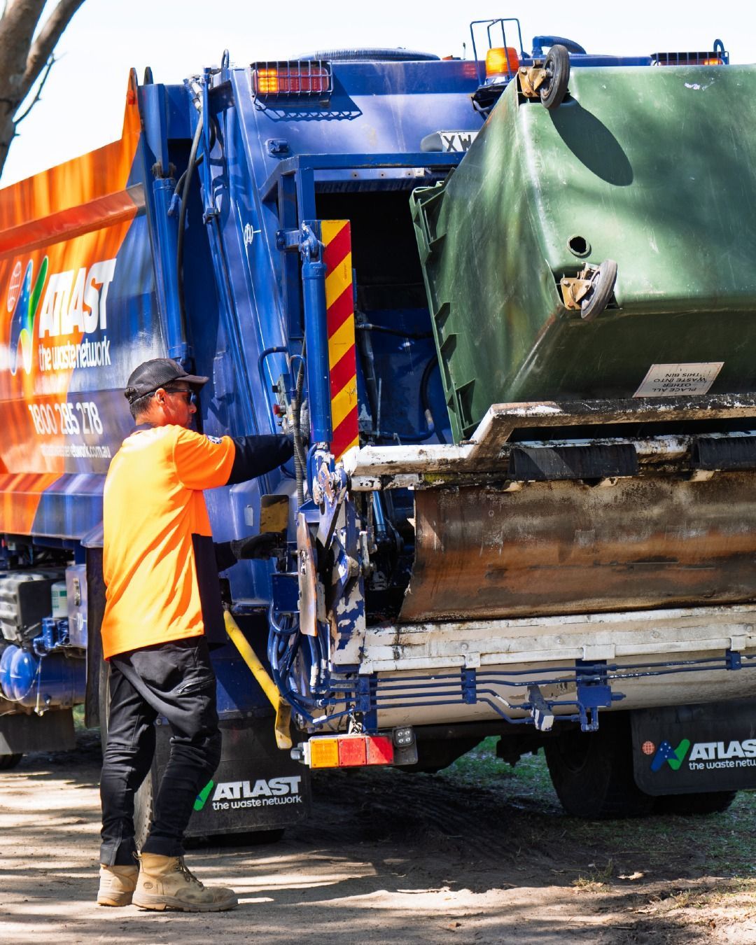 Waste collector emptying a green bin into a blue and orange garbage truck.