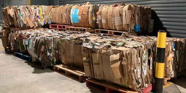 Large stacks of compressed cardboard bales on wooden pallets in a warehouse setting.