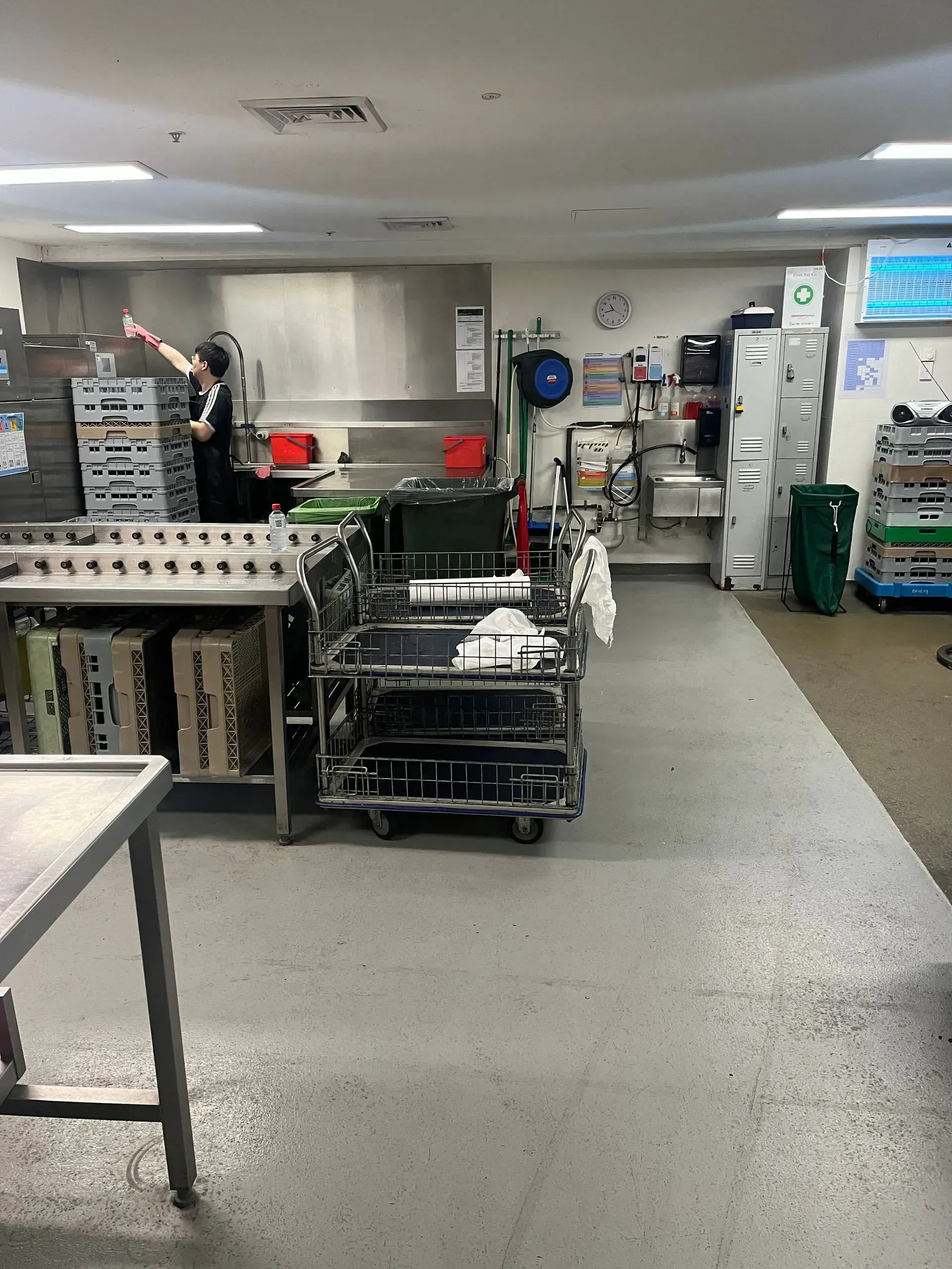 Interior of a sterile kitchen; person working near stacks of metal containers, equipment on rolling carts.