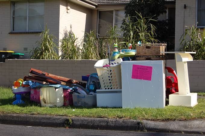 Pile of discarded household items, including a fridge, toys, and wood, on a curb near a residential building.