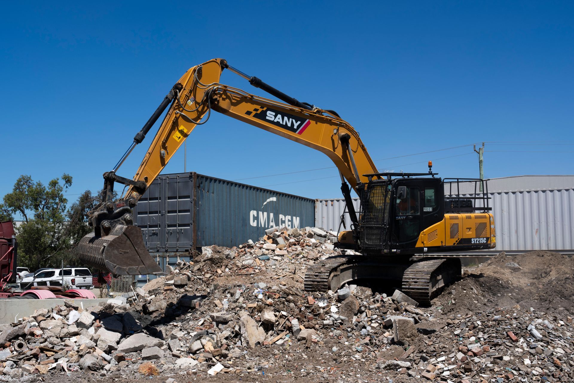 Yellow excavator scooping up debris near shipping containers under a blue sky.