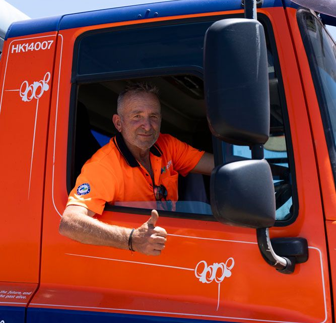 Man in orange shirt gives thumbs up from truck cab. Orange and blue truck has logo.