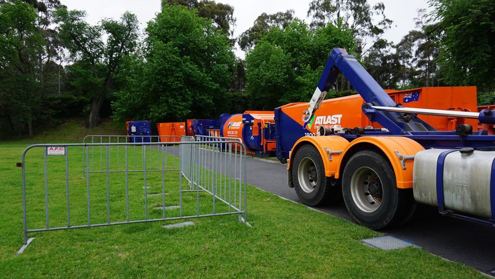Truck and several large orange and blue bins on grass with trees in the background, a small fence in front.