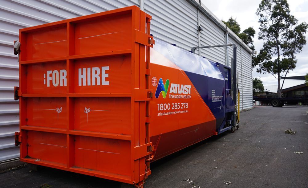 Orange and blue Atlas waste disposal bin for hire, parked near a building.