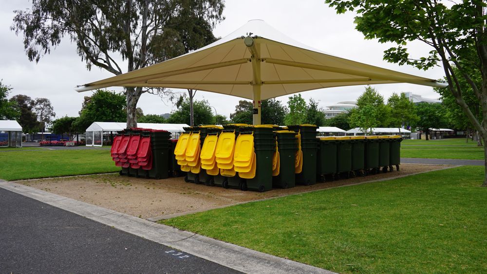 Group of trash cans in various colors under a large, beige umbrella, on a green lawn.