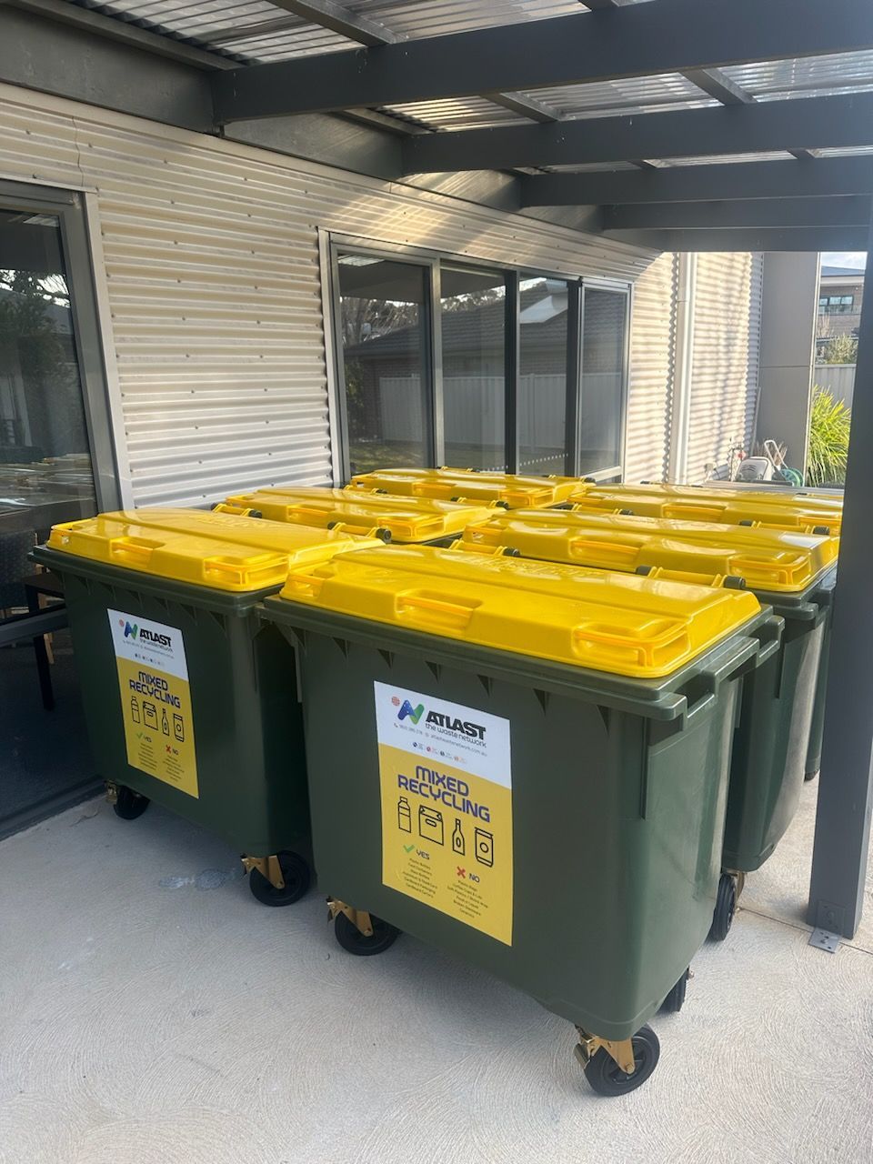 Six green recycling bins with yellow lids under a shelter.