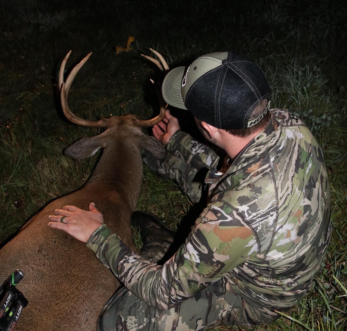 Man in camo touches a dead deer's antlers. Outdoors at night; archery bow visible.