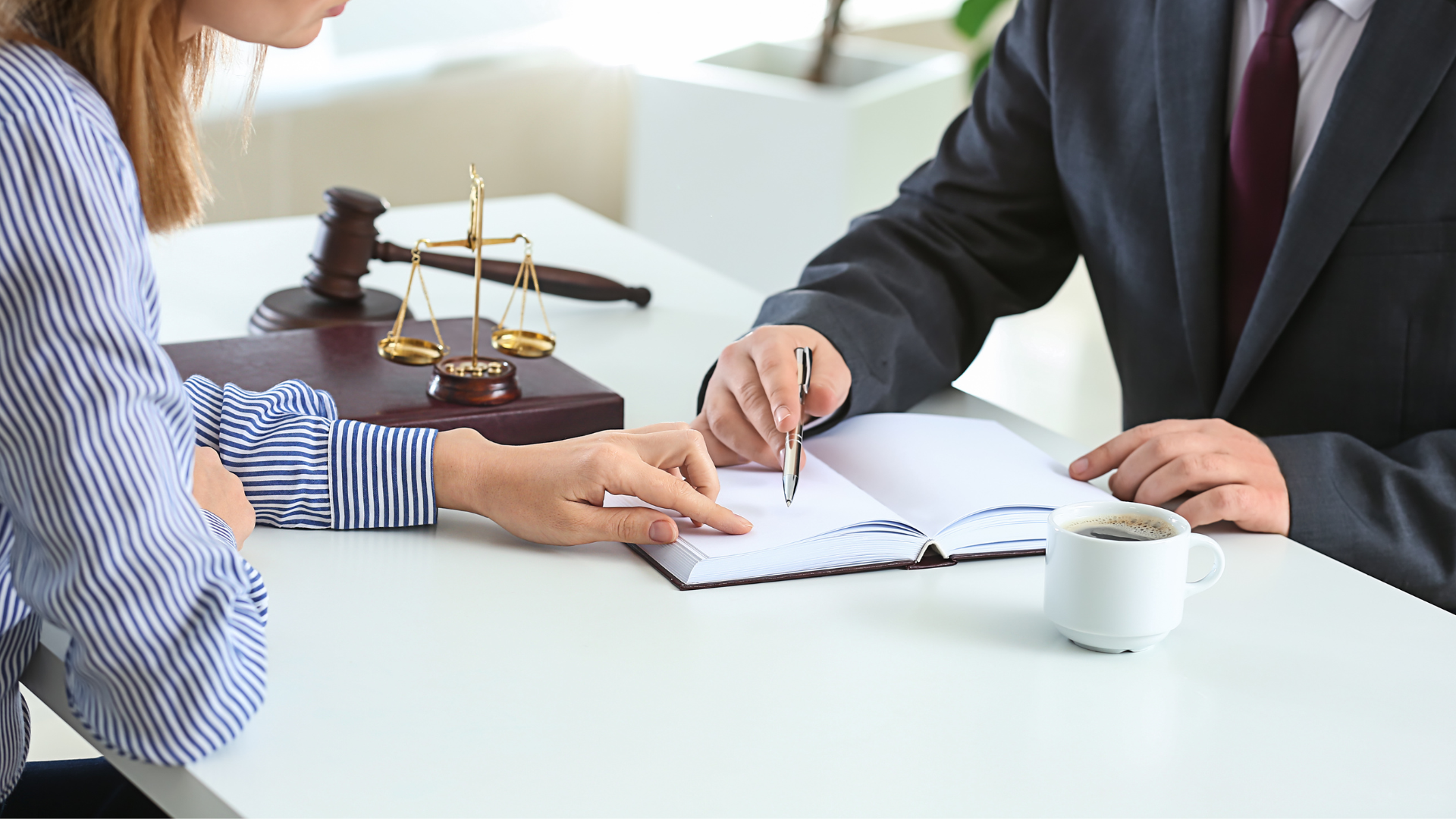 Person pointing at open book with pen, seated at a desk with another person. Gavel and scales of justice in background.