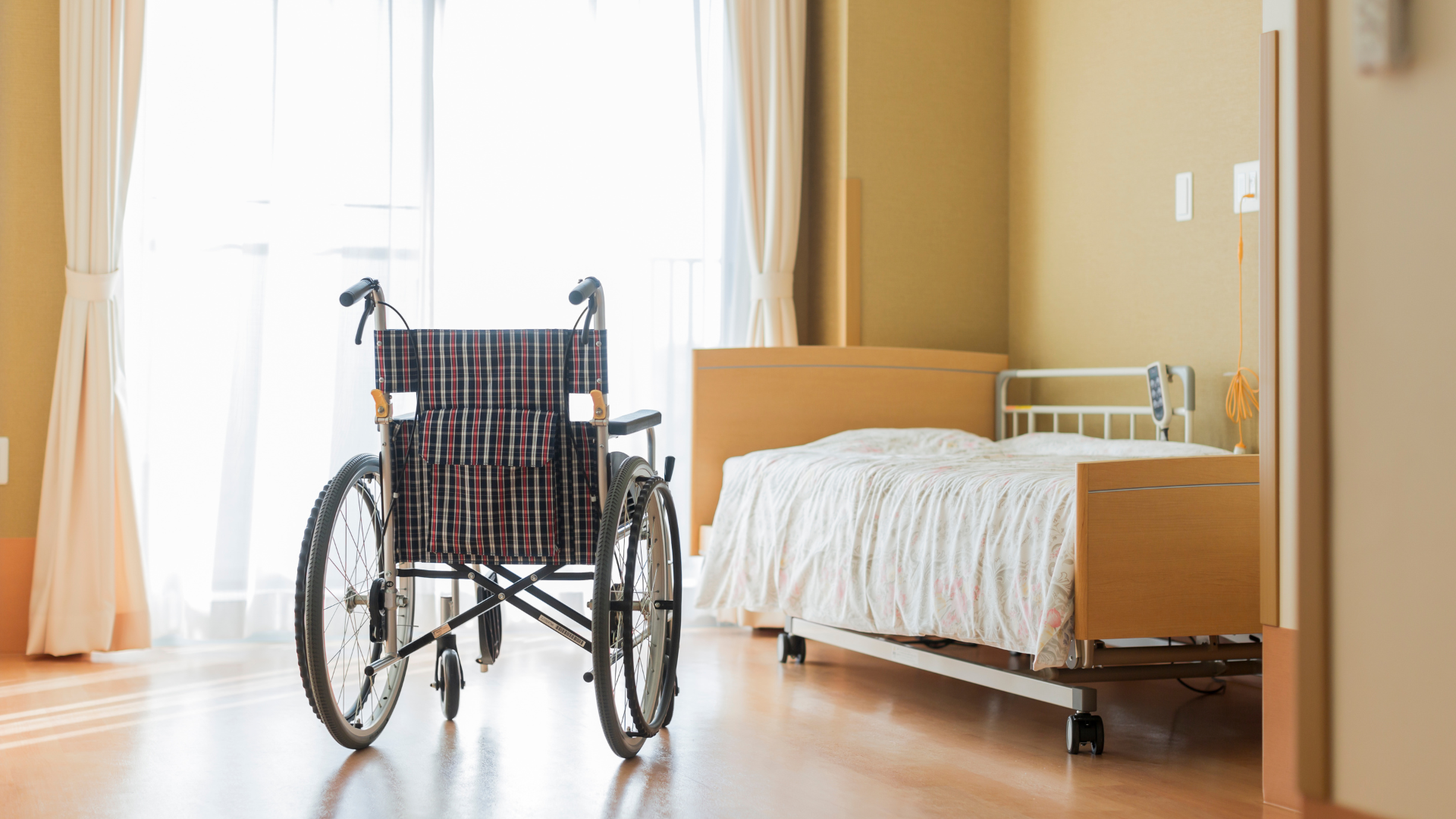 Wheelchair in a bright room with a bed near a window, suggesting a medical or care setting.