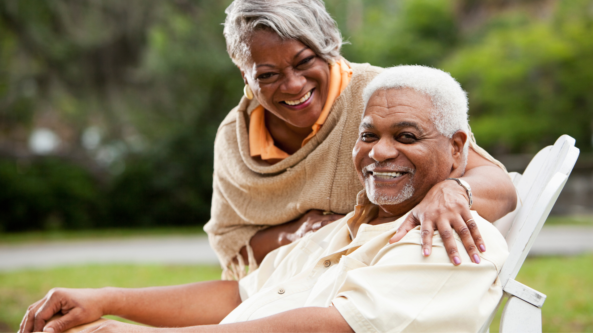 Smiling older couple outdoors, woman's hand on man's shoulder. Man in chair. Green background.