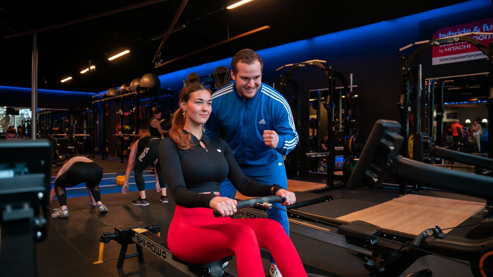 Vrouw roeit met trainer in een sportschool. Vrouw in rode legging en zwart shirt, trainer in blauw Adidas-trainingspak.