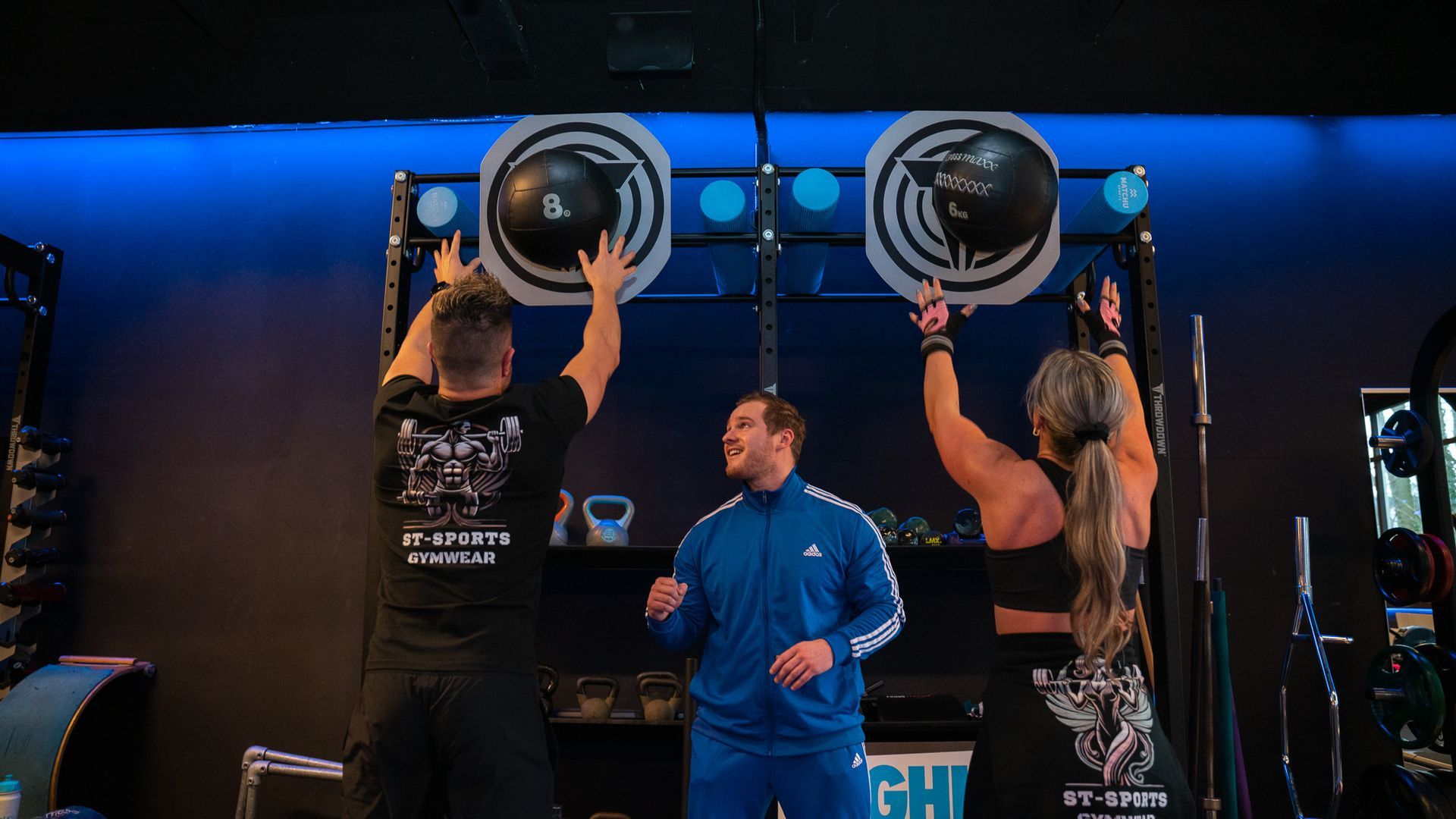 Drie mensen in een sportschool; twee slaan op hangende doelen, één coacht. Blauwe verlichting en apparatuur zichtbaar.