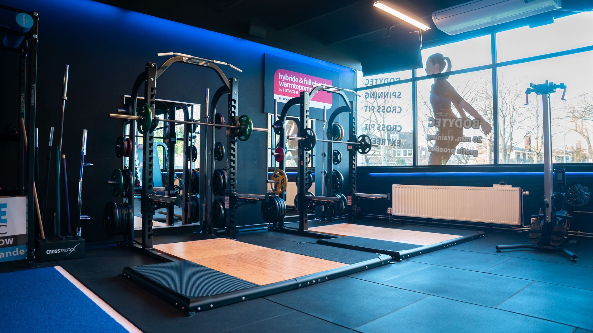 Interieur van een sportschool met gewichthefrekken, blauwe accenten en een groot raam met daarop een persoon.