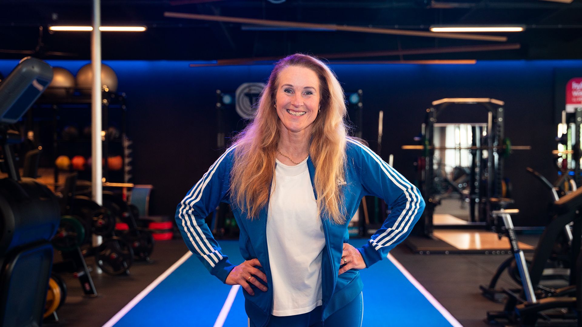 Vrouw in een blauw trainingspak die lacht in een sportschool, handen in haar zij, fitnessapparatuur op de achtergrond.