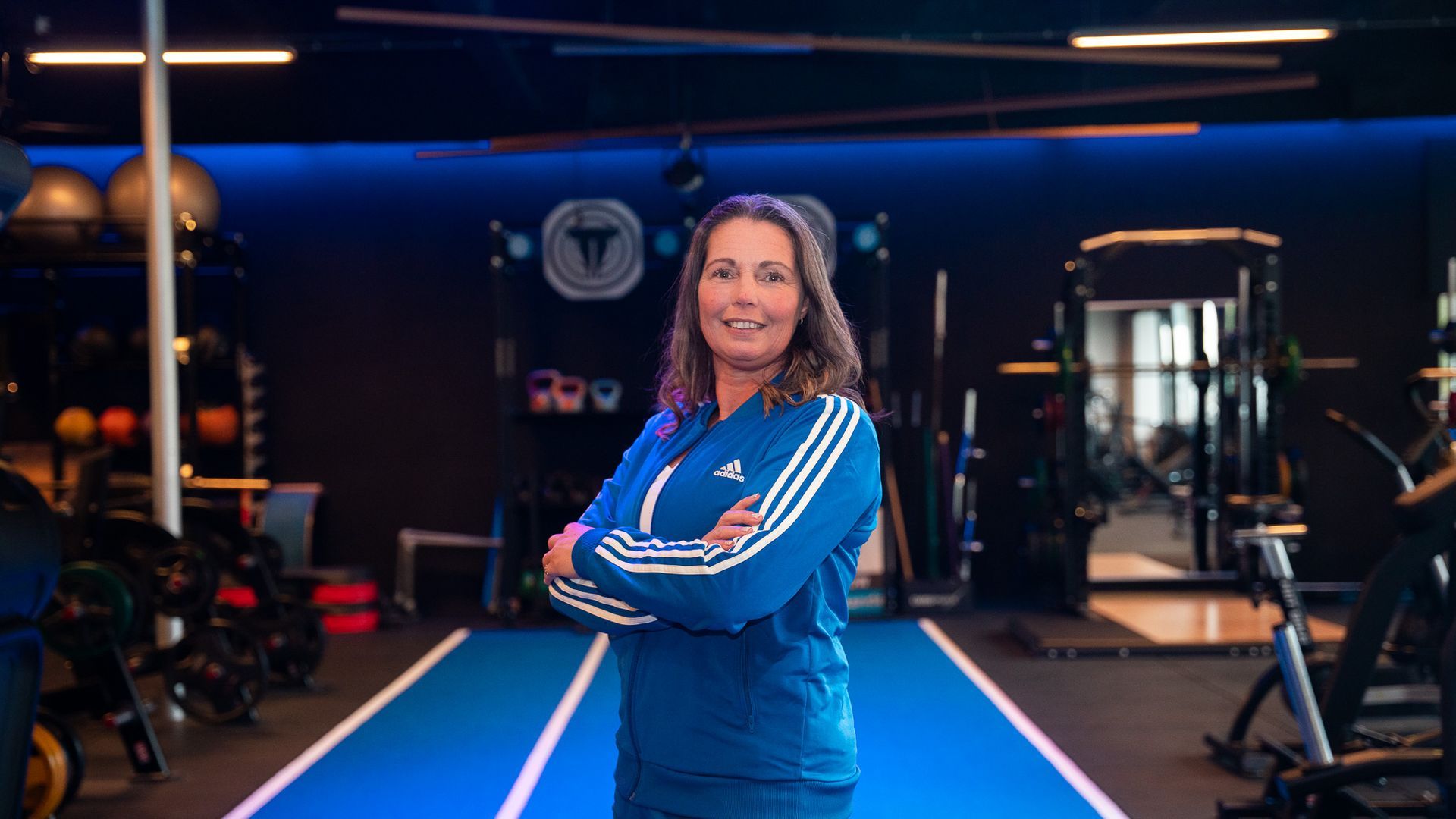 Een vrouw in een blauw trainingspak staat in een sportschool, met haar armen over elkaar en lachend.