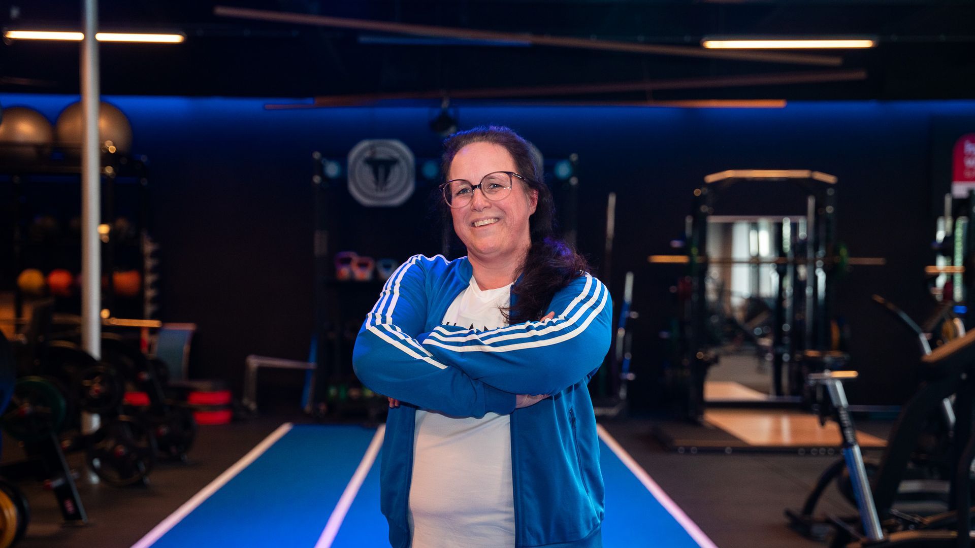 Vrouw in blauw trainingsjack, armen over elkaar, lachend in de sportschool.