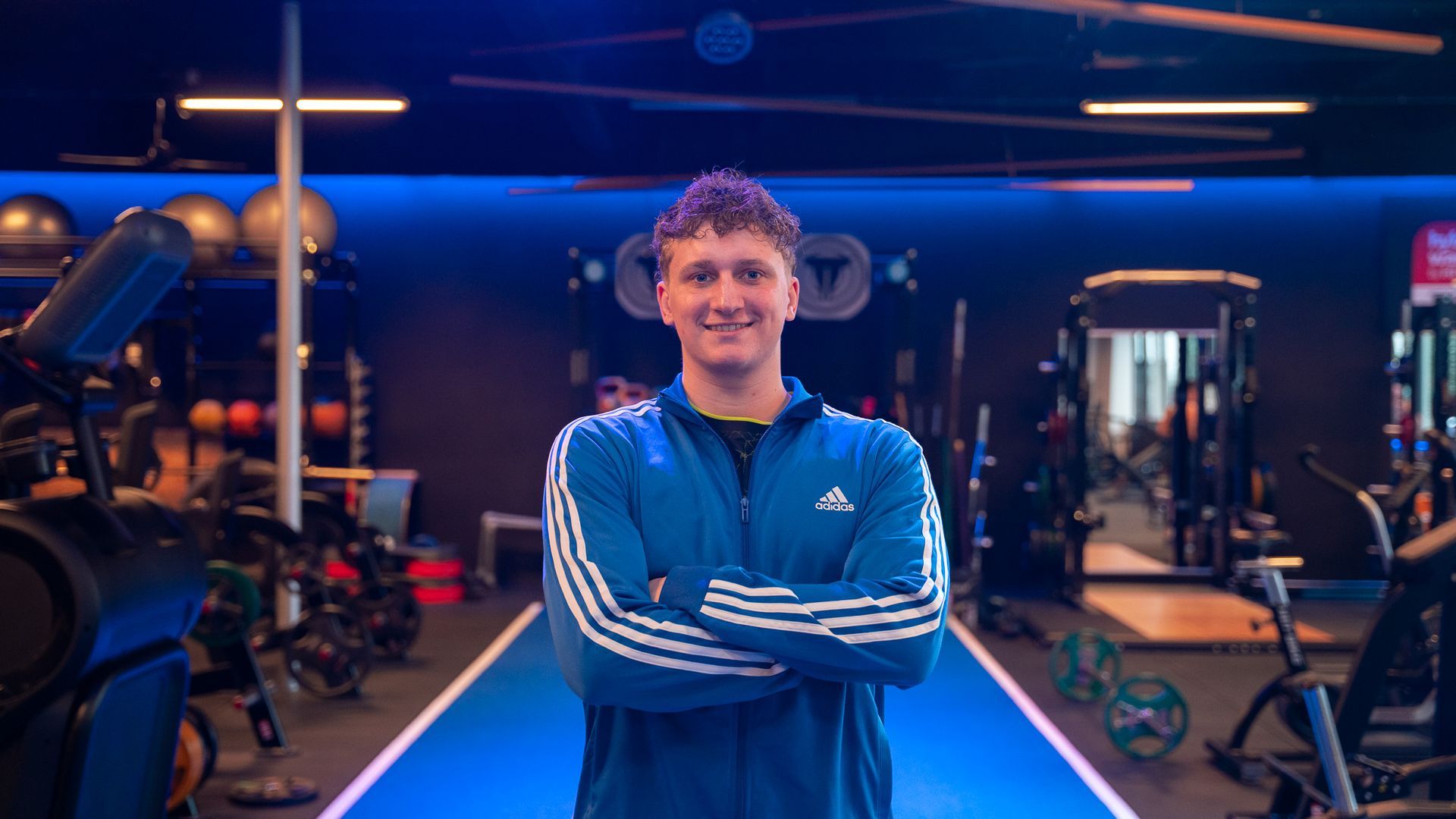 Man met gekruiste armen in een sportschool, gekleed in een blauw Adidas-jack, met een blauw trainingsgedeelte.