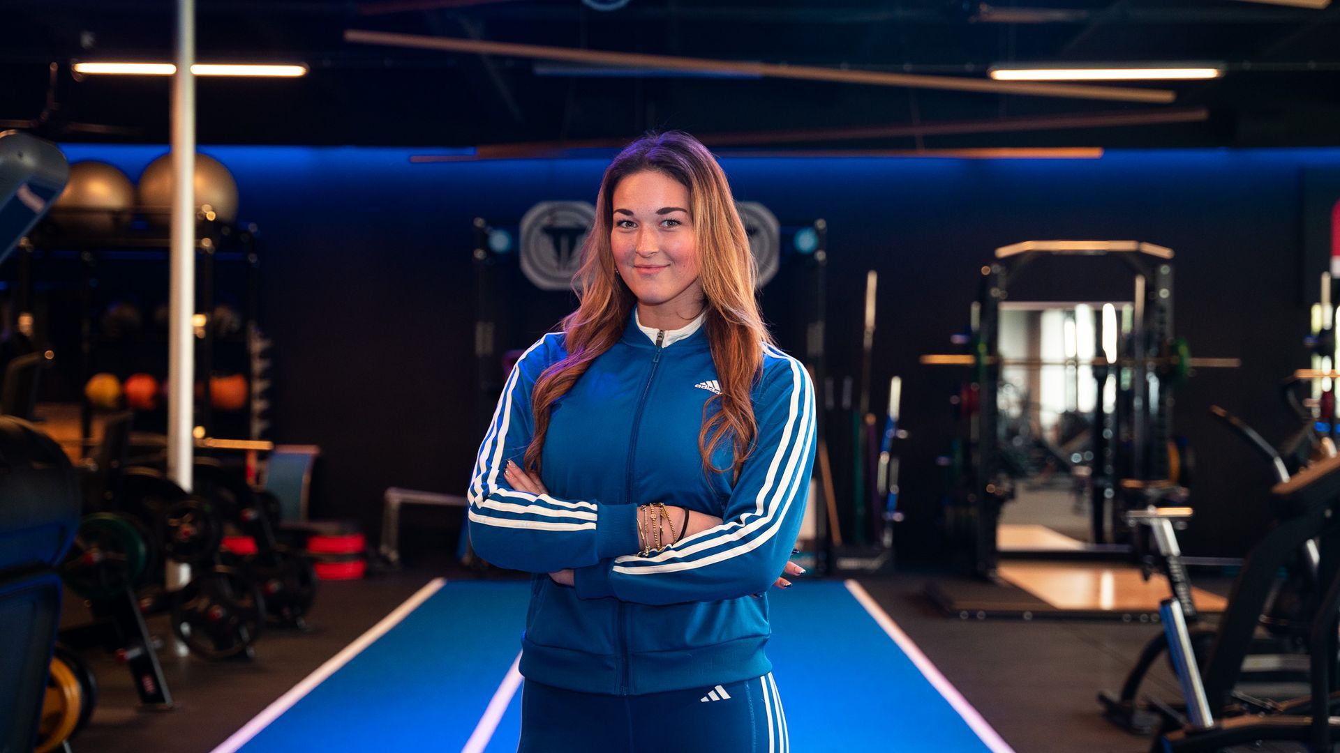 Vrouw in een blauw Adidas-trainingspak, met gekruiste armen, staand in een sportschool.