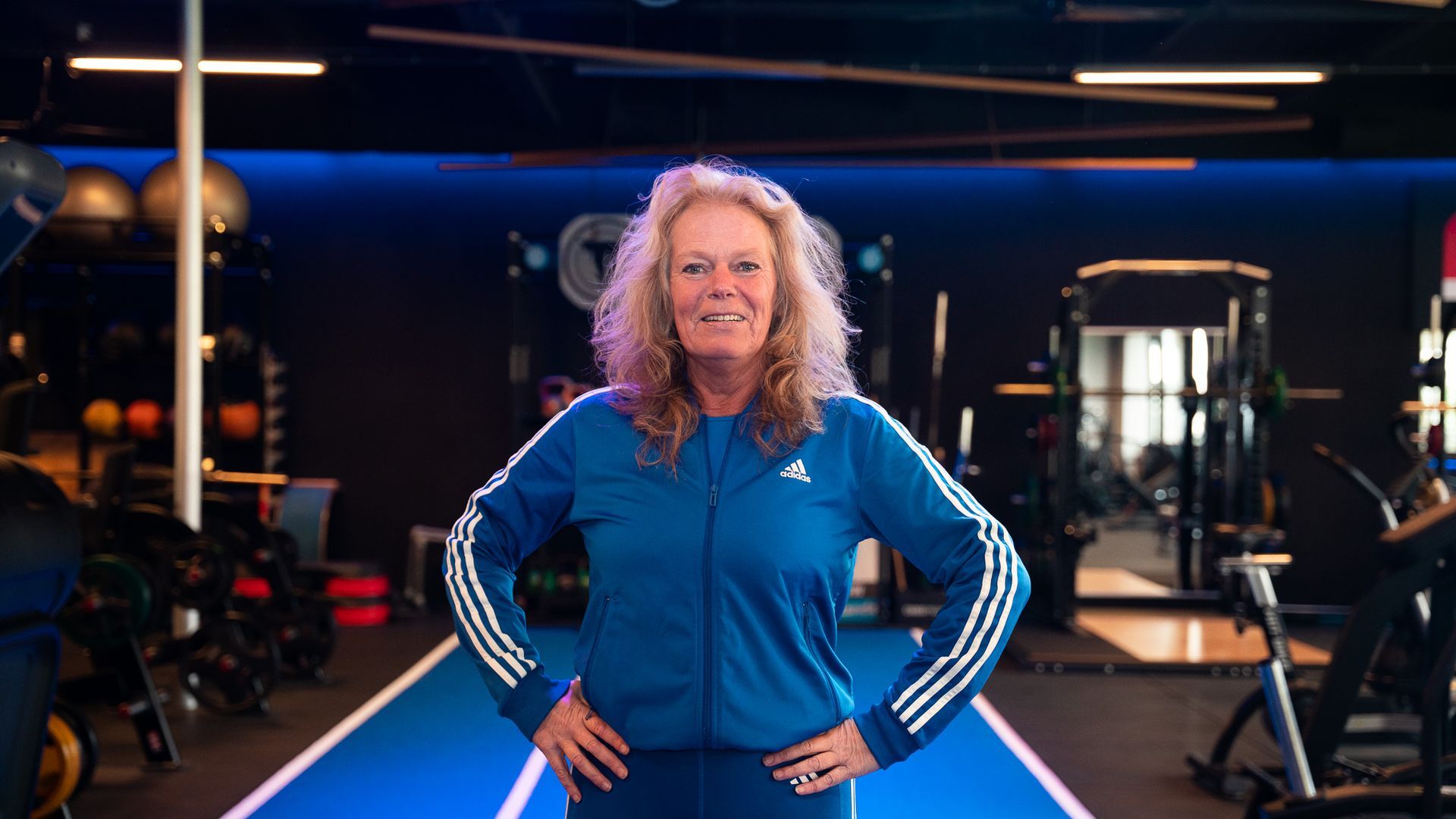 Vrouw in een blauw trainingspak, handen in haar zij, staat in een sportschool.