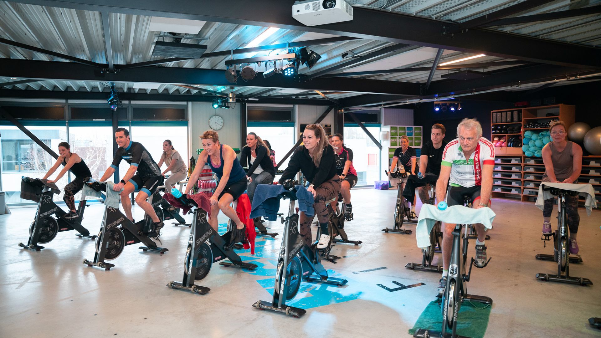 Mensen fietsen tijdens een spinningles. De ruimte heeft een zwart plafond en blauwe verlichting.