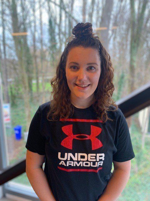 Vrouw met krullend haar in een knot, gekleed in een zwart Under Armour-shirt, glimlachend voor een raam.