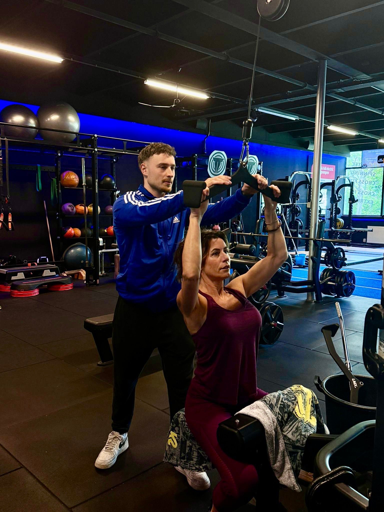Vrouw roeit met trainer in de sportschool; vrouw in rode broek, man in blauw trainingspak.