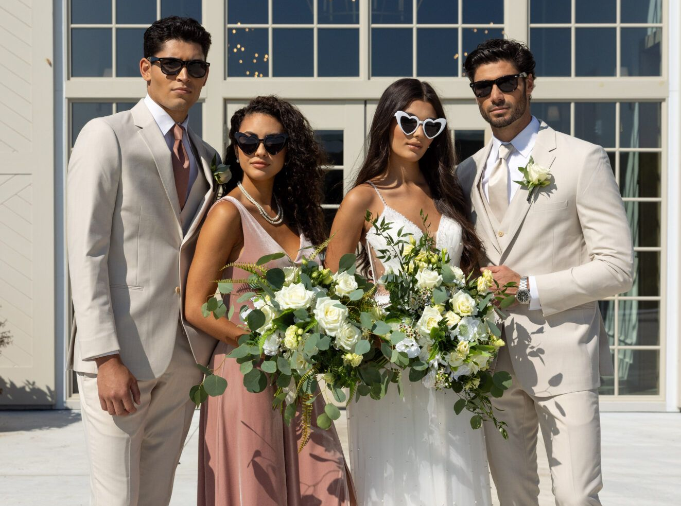 Wedding party posing: bride, bridesmaid, groomsmen in front of building with large windows; beige suits, pink dress, sunglasses.