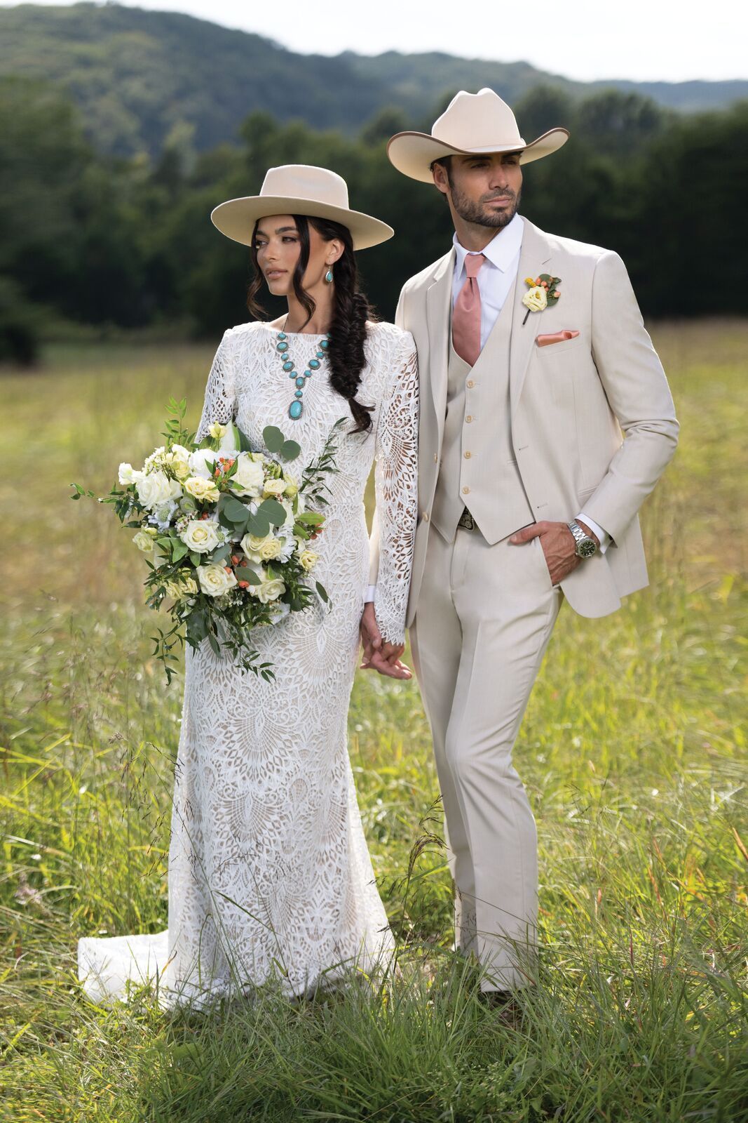 Couple in cowboy hats, standing in a meadow. Bride in lace dress holds bouquet, groom in suit.