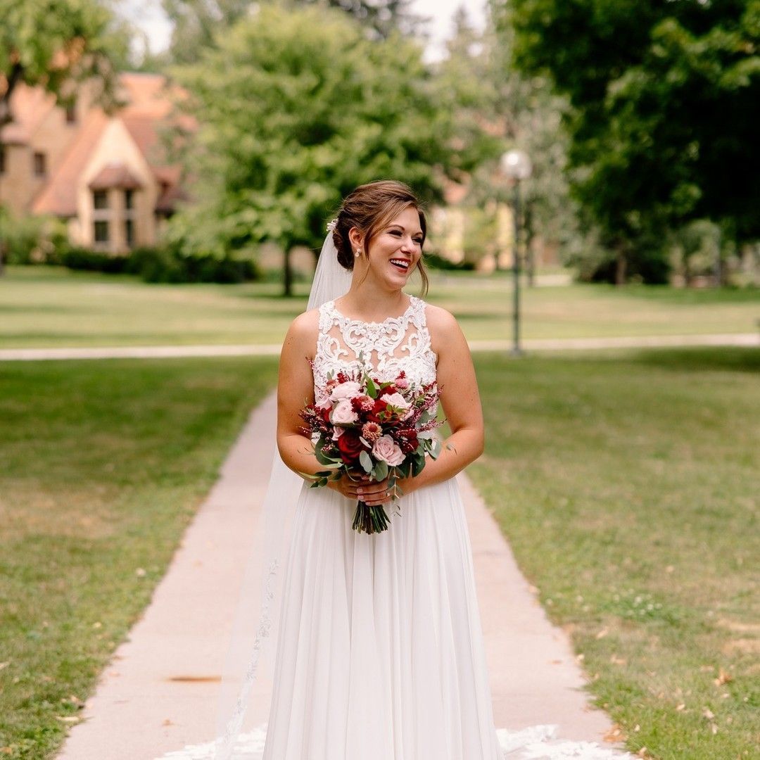 Bride in white wedding dress, smiling, holding bouquet, on a path in front of a building and trees.