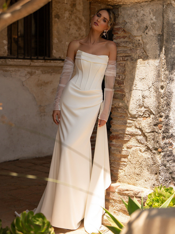 Woman in a white strapless wedding dress with sheer sleeves, leaning against a brick wall.