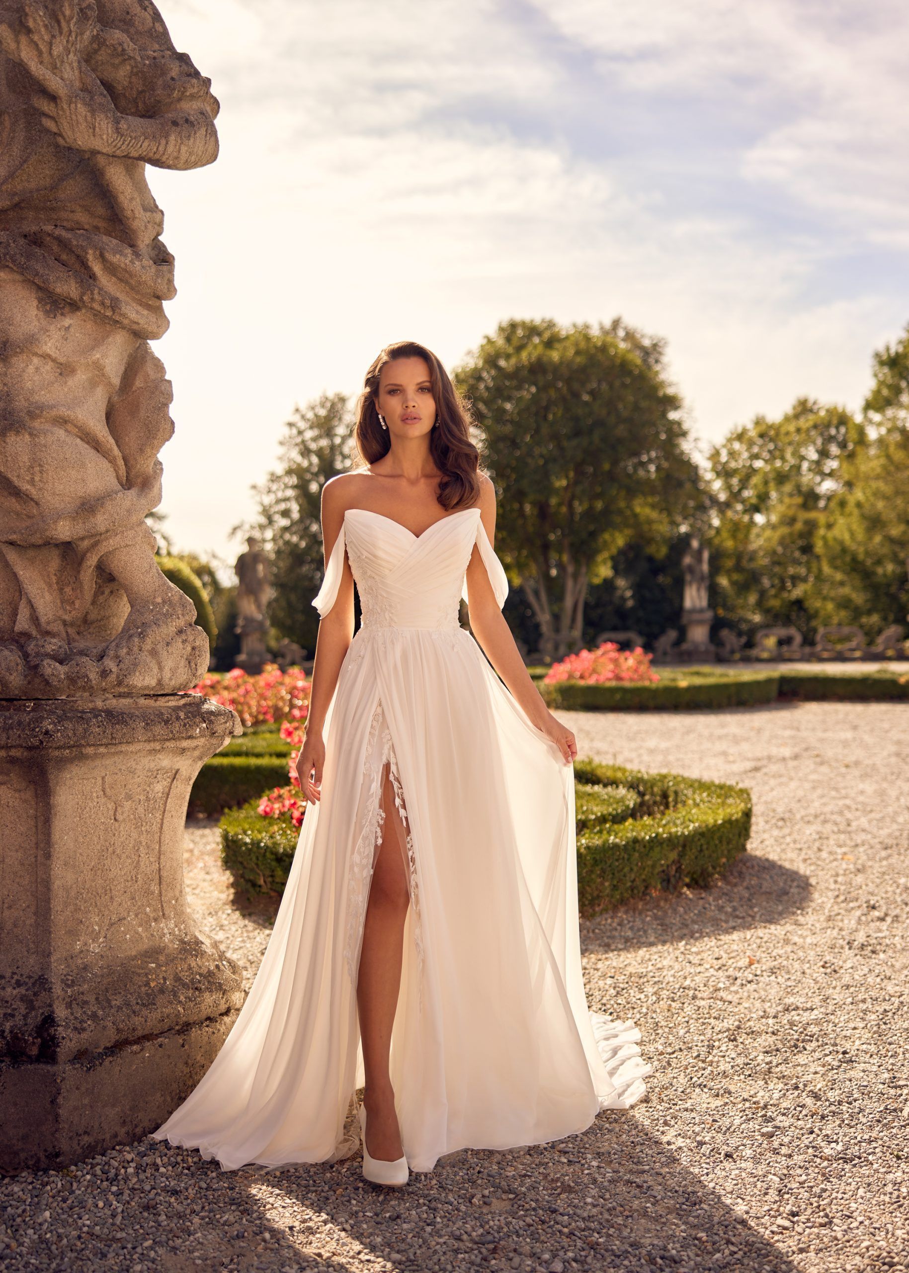 Woman in white off-the-shoulder wedding dress with leg slit, standing outdoors in garden.