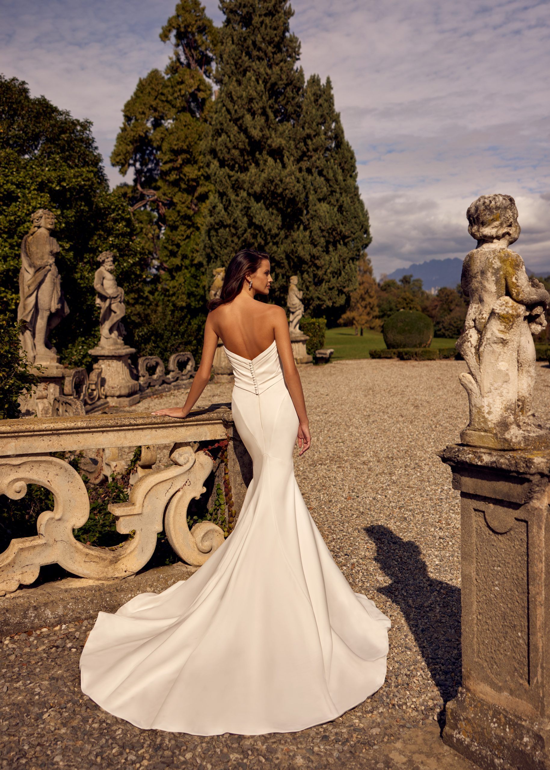 Bride in a white strapless gown with a long train, leaning on a stone balustrade in a garden.