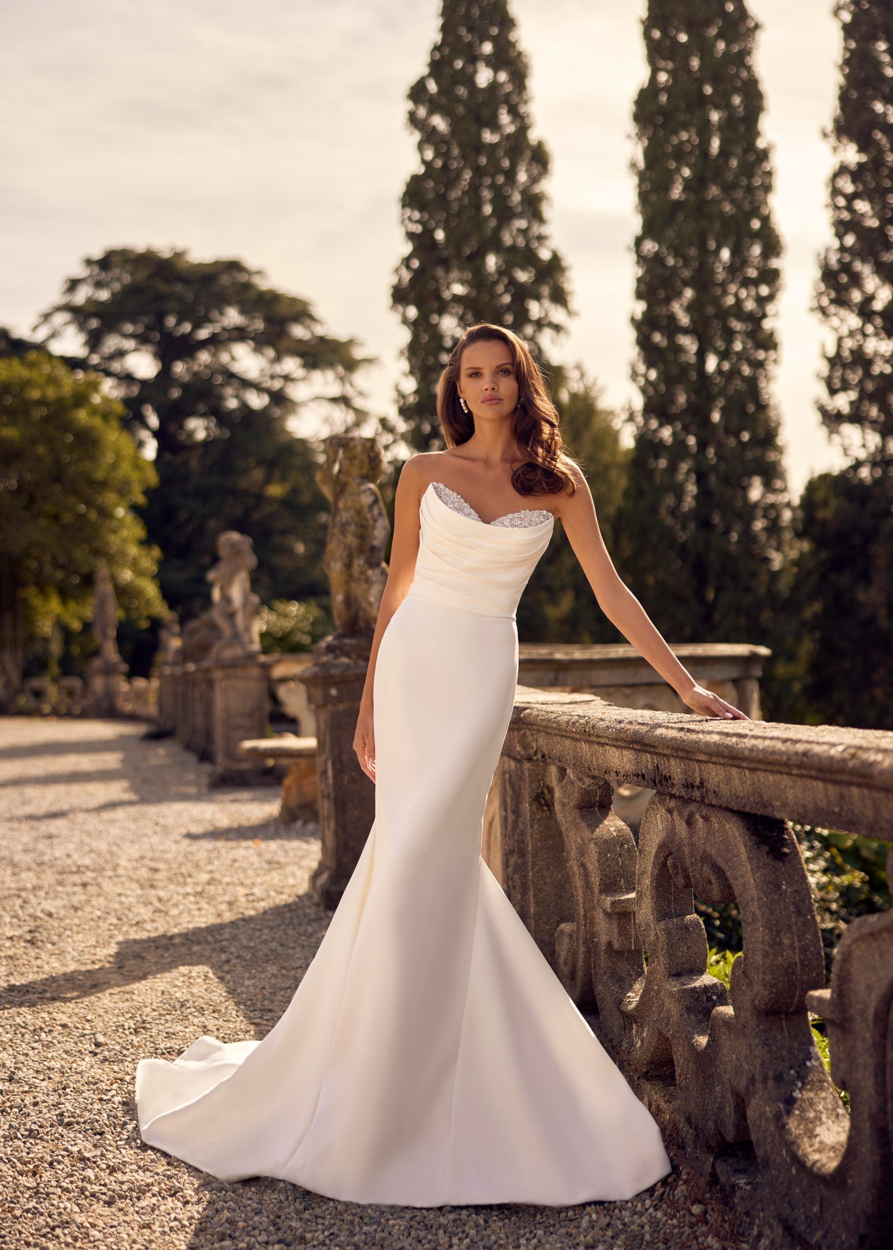 Woman in a white mermaid wedding dress poses on a stone balcony, with tall trees in the background.