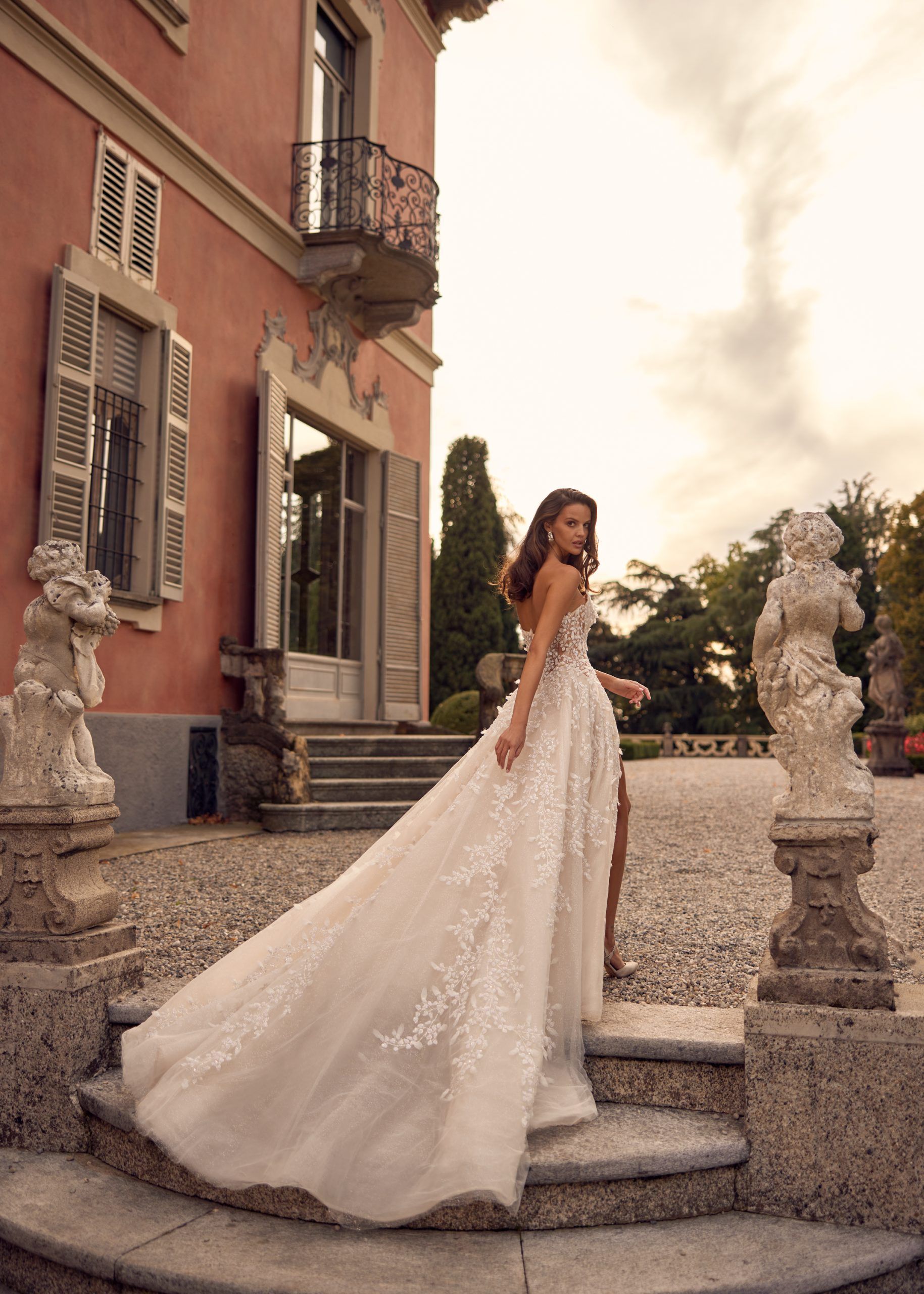 Woman in wedding dress on steps of a villa, looking over shoulder, white gown with floral details, long train.