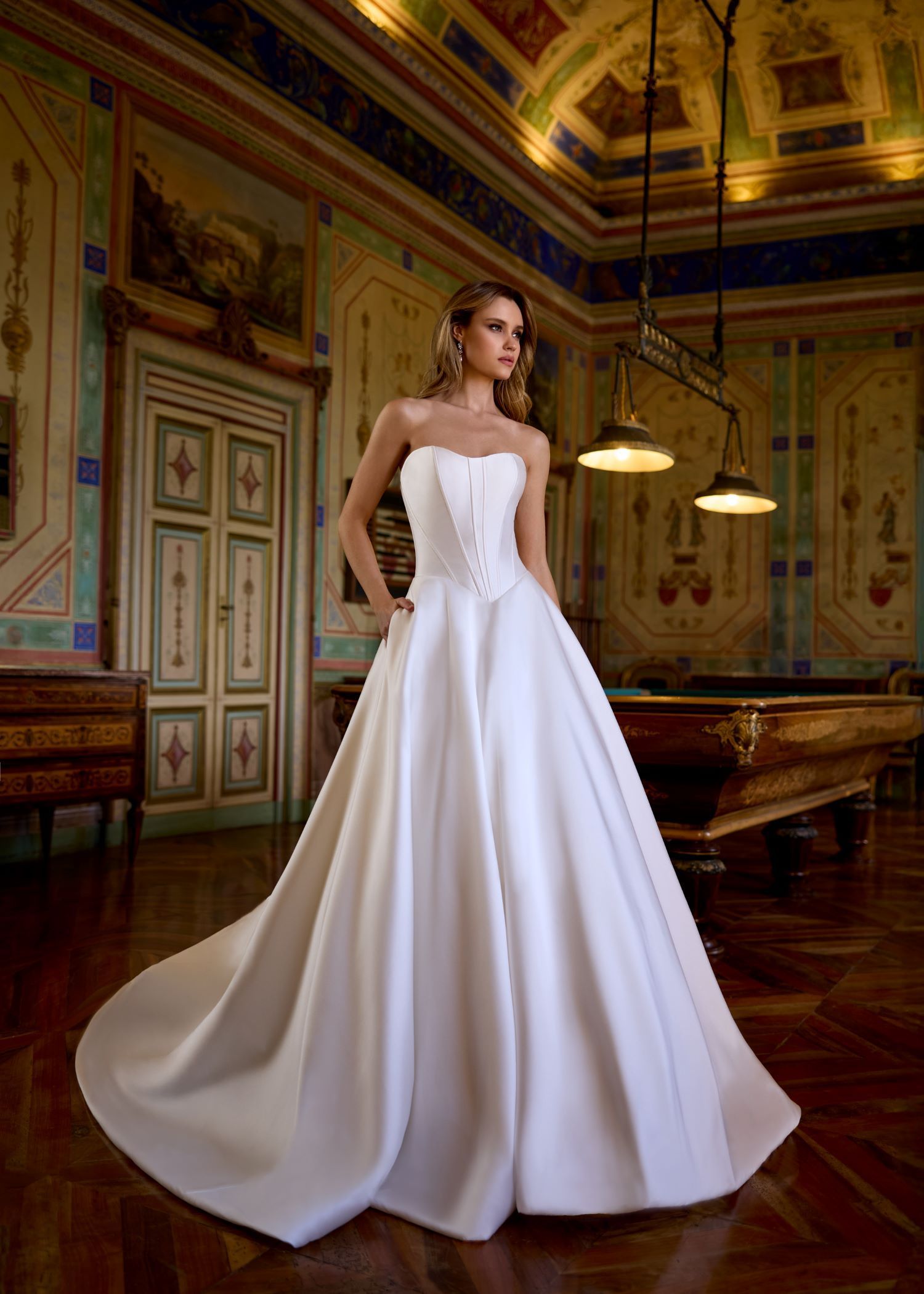Bride in strapless white satin gown, standing in a room with a pool table and ornate ceiling.