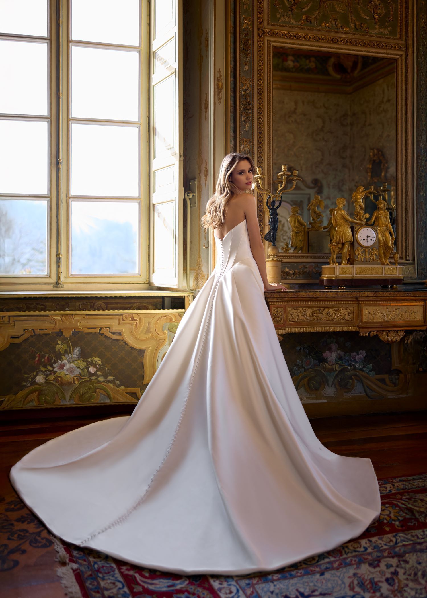 Bride in strapless white gown, posing in front of a window. Elegant interior setting, with gold accents.