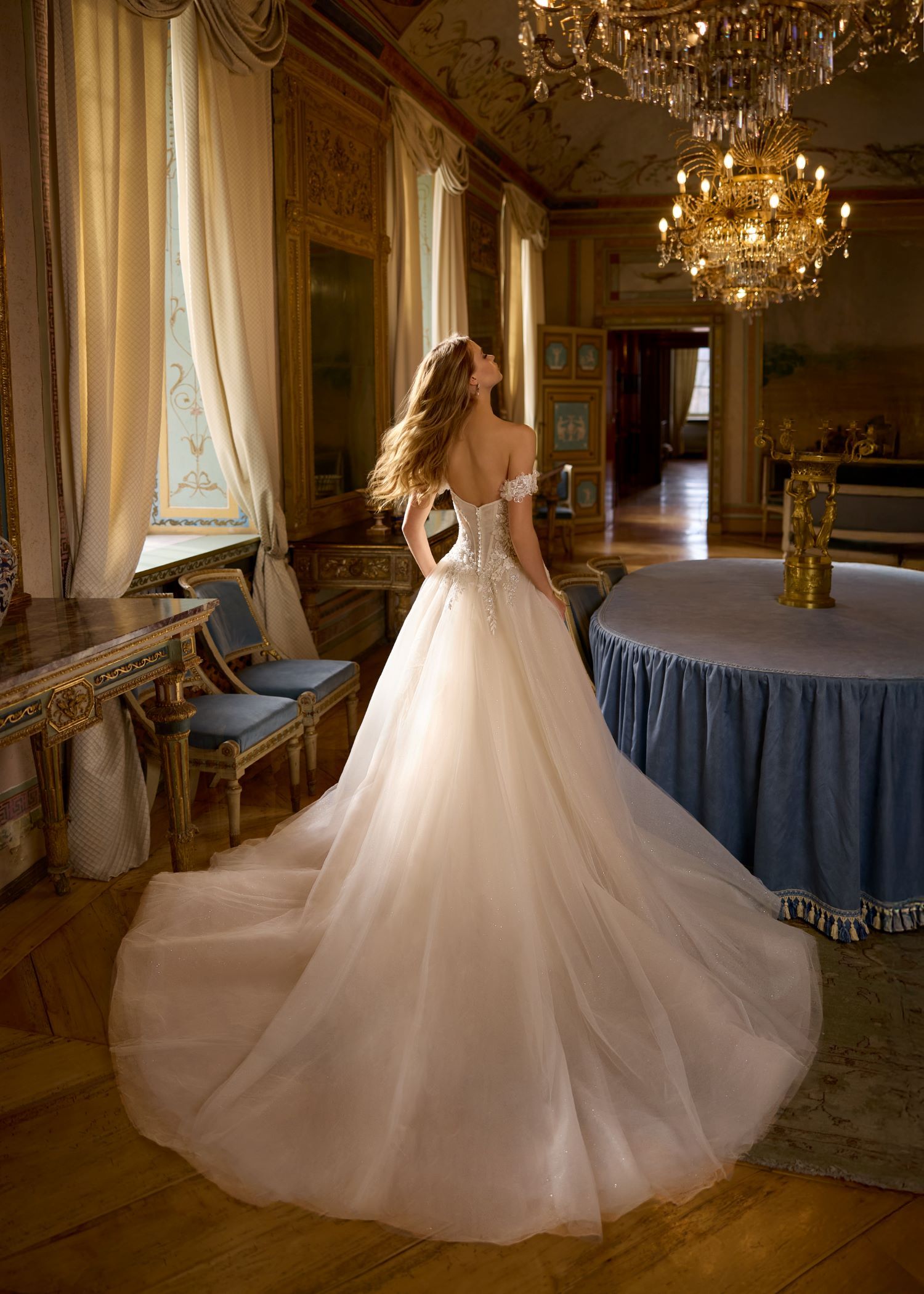 Bride in a white gown with long train, looking up in an ornate room, with a chandelier.