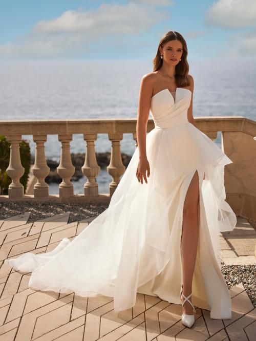 Woman in strapless wedding dress with high slit, standing on a balcony overlooking the ocean.