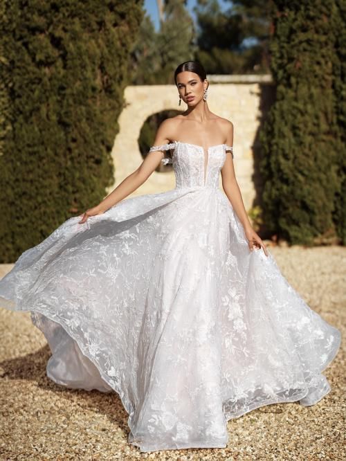 Woman in white off-shoulder wedding dress, arms outstretched, outdoors.