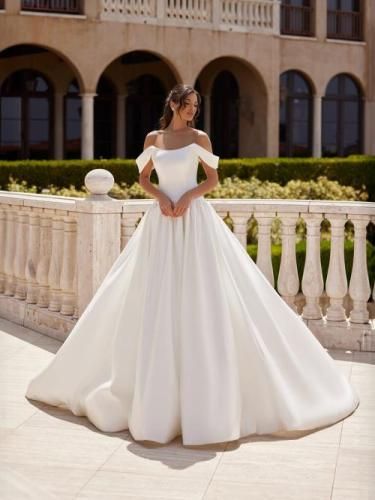 Woman in white off-shoulder wedding dress, standing outdoors, smiling, near stone balustrade and building.