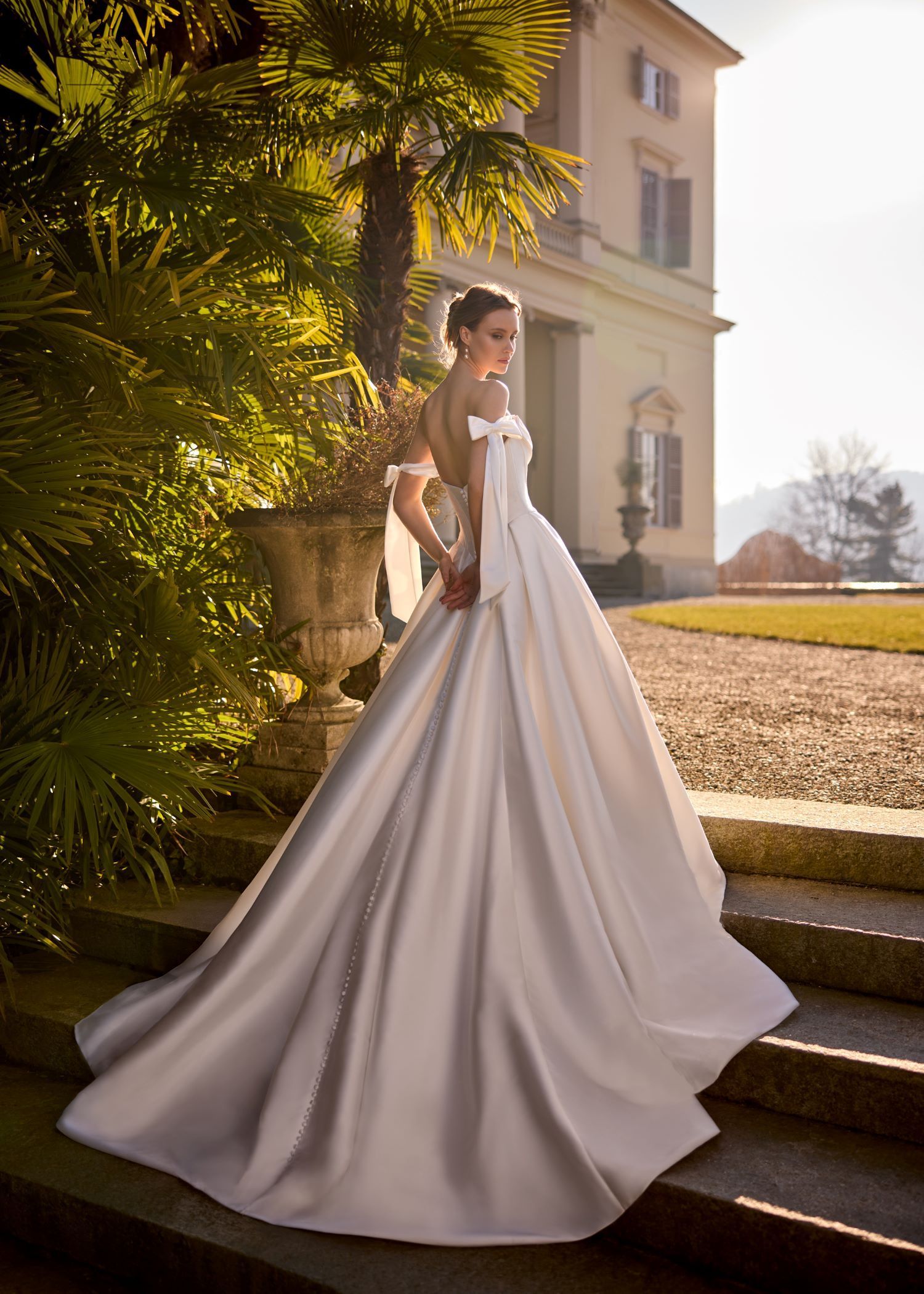 Bride in satin wedding dress stands on steps outdoors, near a building and greenery.